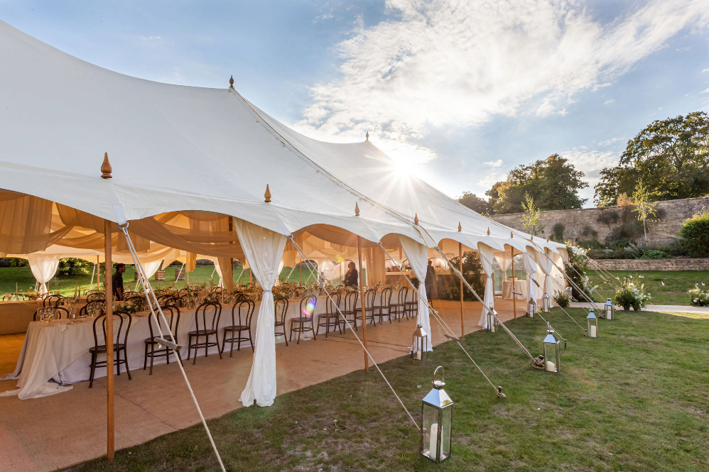 luxury marquee hire for outdoor weddings. Marquee in a garden with open sides for lovely views. Inside long tables are set up for a banquet. The sin shines and catches the beautiful opaque drapes inside the tent