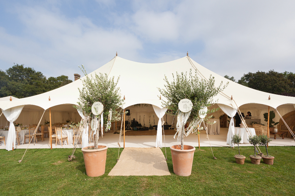 Wedding marquee for an outdoor reception in a garden decorated with greenery and olive trees in terracotta pots. The marquee has open sides so you can see the round tables and wooden dance floor inside